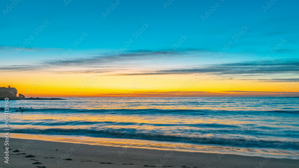Obraz premium Sunrise view on the beach in Sydney, Australia. In the background, the sky with the sun rising with orange and blue colors, while it contrasts with the sea and the silhouettes of people bathing. 