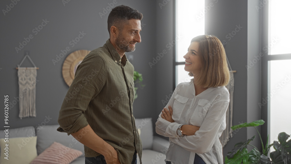 Fototapeta premium Man and woman standing together in a cozy living room, smiling and looking at each other, creating a warm atmosphere of love and togetherness in their home.