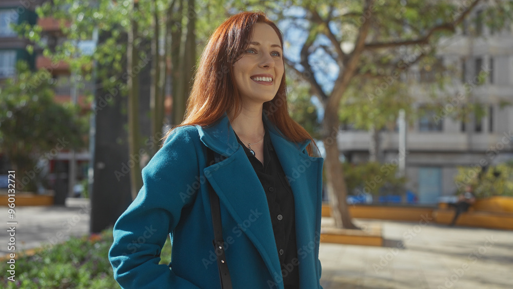Smiling redhead woman in stylish teal coat enjoying a sunny day in a city park with buildings in the background. Smiling redhead woman in stylish teal coat enjoying a sunny day in a city park with buildings in the background.