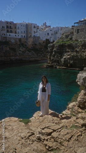 A beautiful young hispanic woman stands on a rocky cliff overlooking the turquoise sea, with the historic buildings of polignano a mare, puglia, italy in the background.