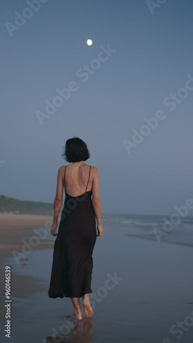 A young woman with short hair, wearing a long flowing dark dress, walks barefoot on the beach as the moonlight shines on the water behind her.
