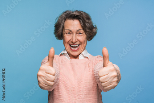 Mature satisfied happy smiling caucasian woman female pensioner in casual clothes showing two thumb up like gesture isolated on blue color background studio portrait. People lifestyle concept photo