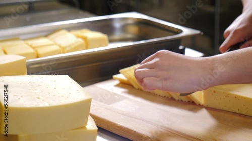 Worker is using a knife to slice a block of pale yellow cheese with small holes on a wooden cutting board. 4 k video