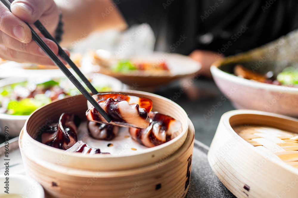 A close-up of a hand using chopsticks to pick up a piece of dim sum from a bamboo steamer, with more dim sum in the background. The image captures the essence of traditional Asian cuisine and dining. A close-up of a hand using chopsticks to pick up a piece of dim sum from a bamboo steamer, with more dim sum in the background. The image captures the essence of traditional Asian cuisine and dining.