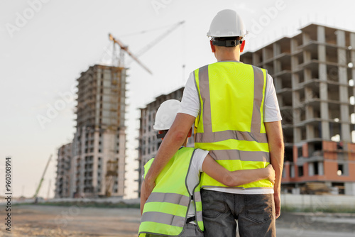 Father and son in protective helmets and vests look at the construction of a building at sunset.