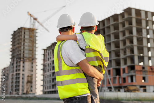 Father and son in protective helmets and vests look at the construction of a building at sunset. photo