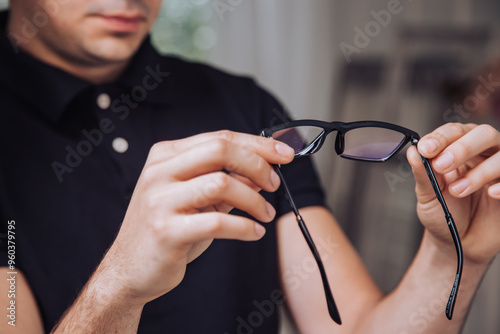 Man inspects black-framed glasses indoors under soft natural light. A man carefully examines a pair of black-framed glasses