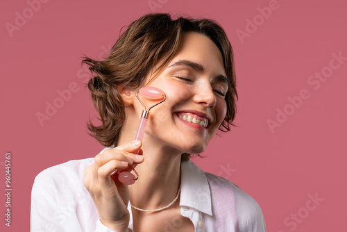 Young woman with rose quartz stone roller on pink background. Facial self care