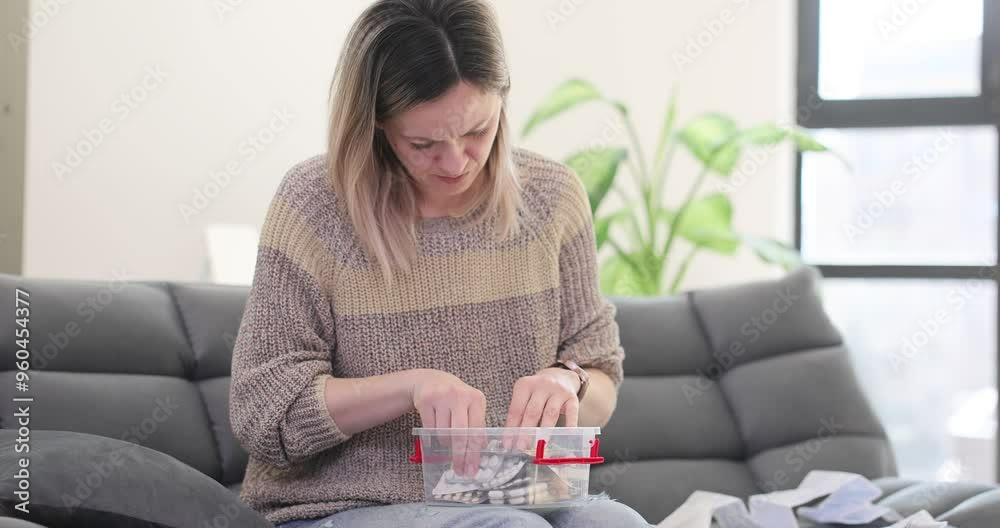 Young woman rummages through home medicine kit sitting on sofa. Lady eyes scanning various pills in search of remedy to address symptoms
