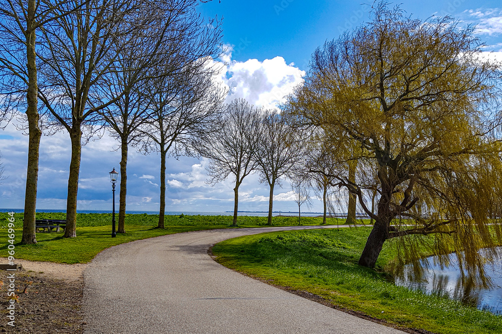 Winding country road between bare trees against blue sky with plenty of white clouds, Ijsselmeer lake in background, sunny spring day in Medemblik, Noord-Holland, Netherlands Winding country road between bare trees against blue sky with plenty of white clouds, Ijsselmeer lake in background, sunny spring day in Medemblik, Noord-Holland, Netherlands