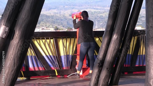 Mujer vista desde atrás usando binoculares públicos en mirador con bandera de Colombia y paisaje de fondo