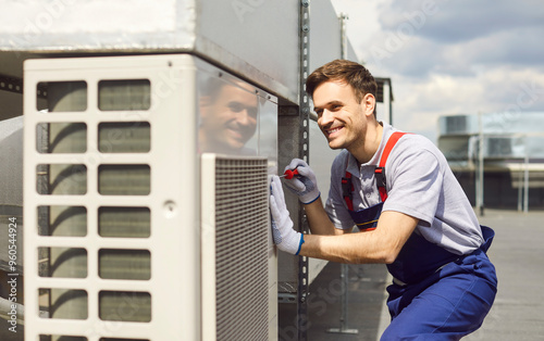 Young professional technician wearing uniform installing and fixing air conditioner using screwdriver sitting on the rooftop. Smiling male repairman repairing ventilation system outdoors. photo