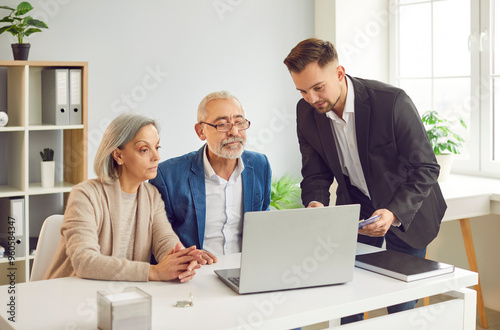 Smiling happy elderly couple talking with man advisor about health insurance or investments sitting at the desk in office with laptop. Senior man and woman talking with male financial agent.