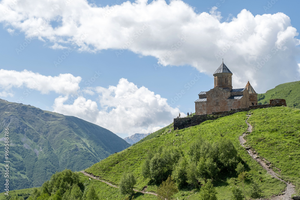 Gergeti Trinity Church overlooks green mountains under a blue sky. The church symbolizes faith in the Caucasus Mountains