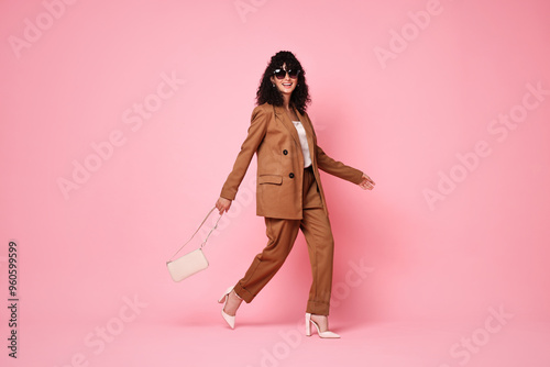 Beautiful young woman in stylish suit and sunglasses with bag walking on pink background