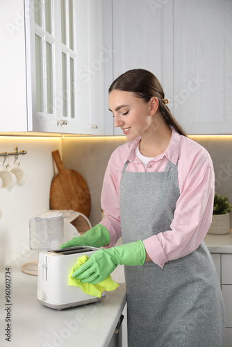 Beautiful young woman wiping toaster with rag at countertop in kitchen photo