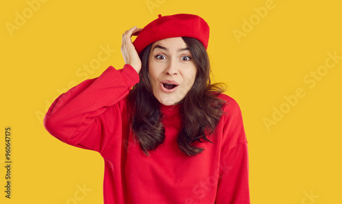 Photo of beautiful impressed young woman touching her head. Surprised brunette girl in red beret and sweatshirt staring at camera with open mouth over isolated on yellow background