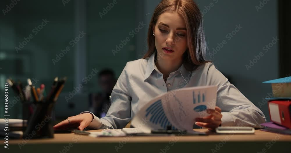 Tired young woman worker examines report with charts. Lady glances at smartphone to check time as working on paperwork late at night in office