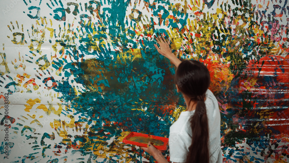 Naklejka premium Young happy girl holding the color tray and paint colorful stained wall to express idea and enhance imagination with hand print. Caucasian girl stamp hand to decorate room in art lesson. Edification.