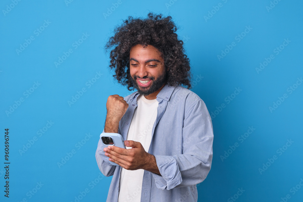 A young man with curly hair expresses excitement as he looks at his phone, smiling broadly. He stands against a bright blue background, radiating happiness in the moment. A young man with curly hair expresses excitement as he looks at his phone, smiling broadly. He stands against a bright blue background, radiating happiness in the moment.