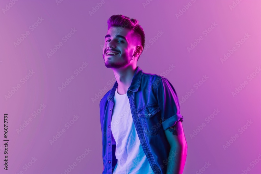 A young man smiles confidently in a denim shirt and white t-shirt while immersed in vibrant neon hues against a purple backdrop