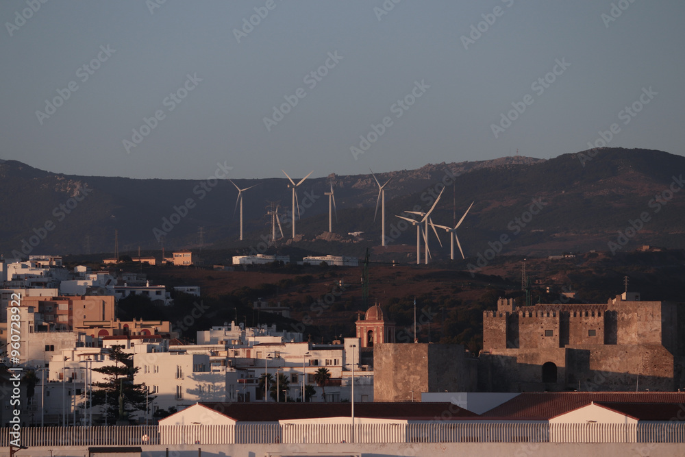 Fototapeta premium View of wind turbines on the mountains with curvy road between them, blue skies with white clouds at sunset with the city of Tarifa in the foreground