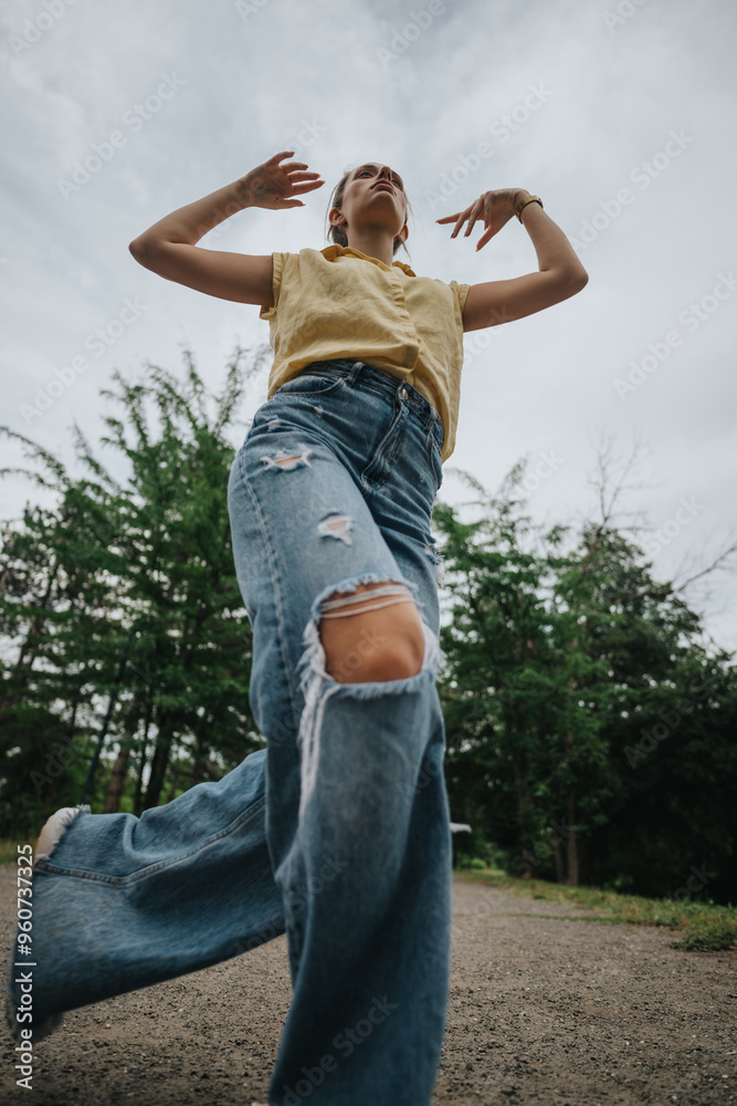 Low angle shot of a woman in ripped jeans and a yellow shirt walking on a path. Green trees in the background.