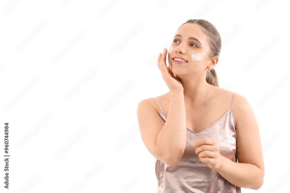 Beautiful young woman applying cosmetic cream on her face against white background
