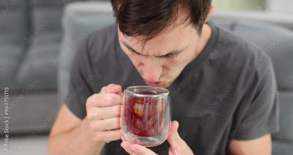 Man holds transparent mug with hibiscus tea sitting on sofa in living room. Male enjoys healthy hot beverage with rich taste in morning Man holds transparent mug with hibiscus tea sitting on sofa in living room. Male enjoys healthy hot beverage with rich taste in morning