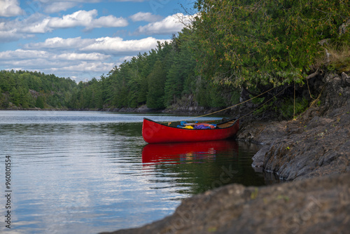 Bright red canoe loaded with camping gear tied to a tree by rugged rocky shore at French River Provincial park, Ontario, Canada. Camping, portaging, adventure, summer activities concept.