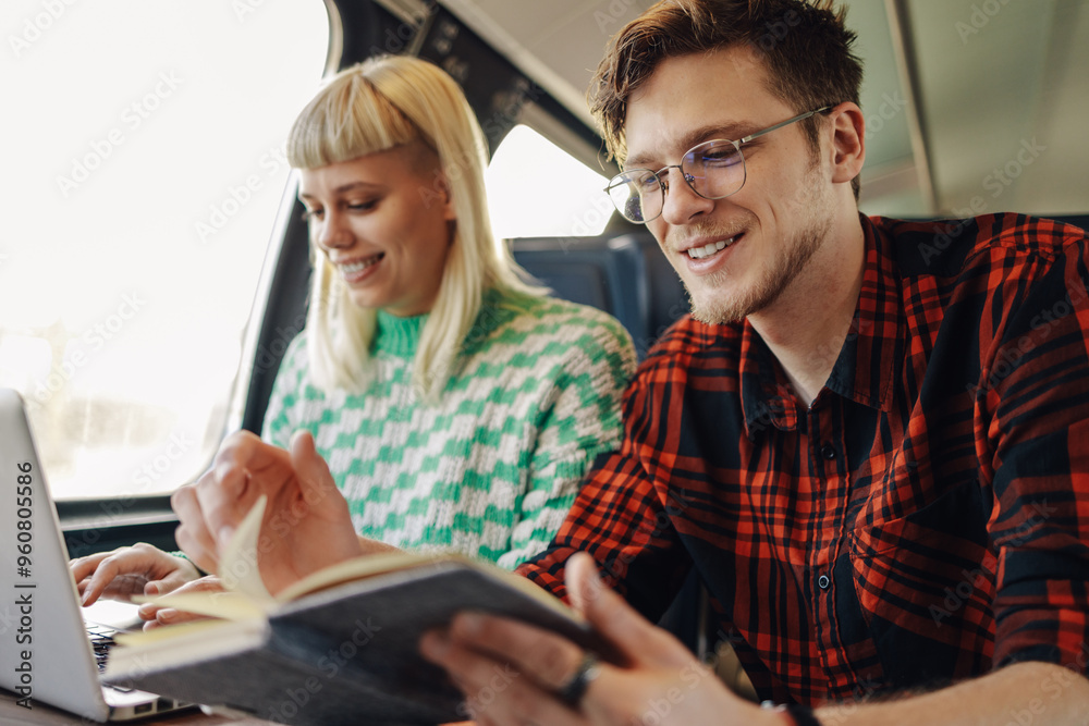Portrait of a happy couple reading a book on a train and using a laptop.
