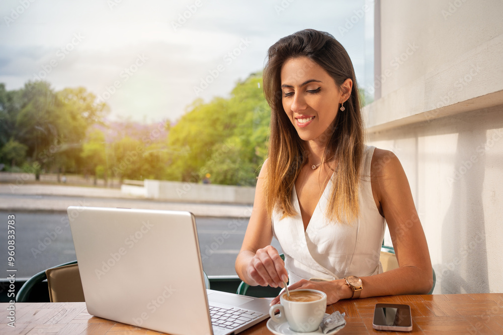 Fototapeta premium Smiling young female freelancer stirring coffee in cup while looking at laptop screen. Beautiful businesswoman sitting in an online meeting while having drink at outdoor cafe.