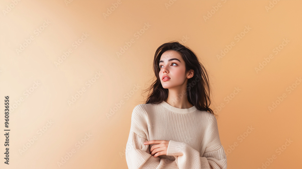 Obraz premium Beautiful girl in a warm jumper against a beige wall points her finger sideways. Dark-haired girl in cosy jumper, copy space on the side