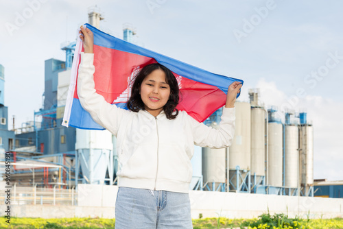 Positive young woman posing with Cambodia flag in hands against background of factory photo