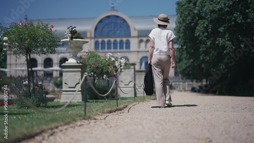 A woman wearing a straw hat is walking in a park walkway at Botanical garden in Colgone, Germany. The park is filled with trees and flowers, and there are several people walking around photo