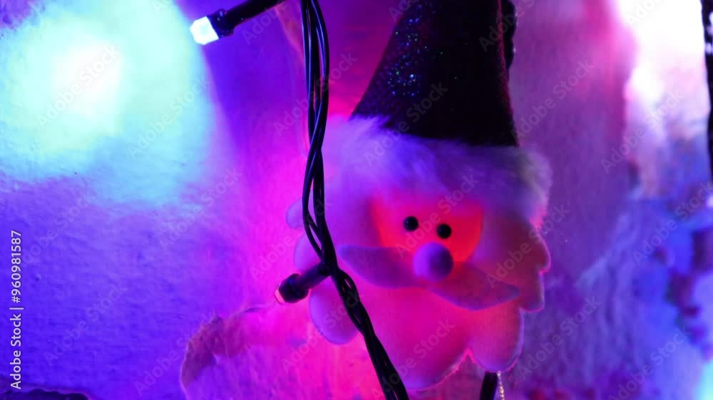 Una toma de cerca de una decoración de navidad y fin de año con una cara de santa claus  rodeado de varias luces con bombillos de varios colores en movimiento en la oscuridad colocadas en una pared 