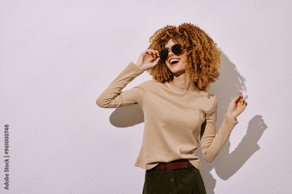 Stylish woman with curly hair wearing sunglasses, posing against a plain light background, showcasing a fashionable look and a cheerful demeanor, perfect for lifestyle concepts Stylish woman with curly hair wearing sunglasses, posing against a plain light background, showcasing a fashionable look and a cheerful demeanor, perfect for lifestyle concepts