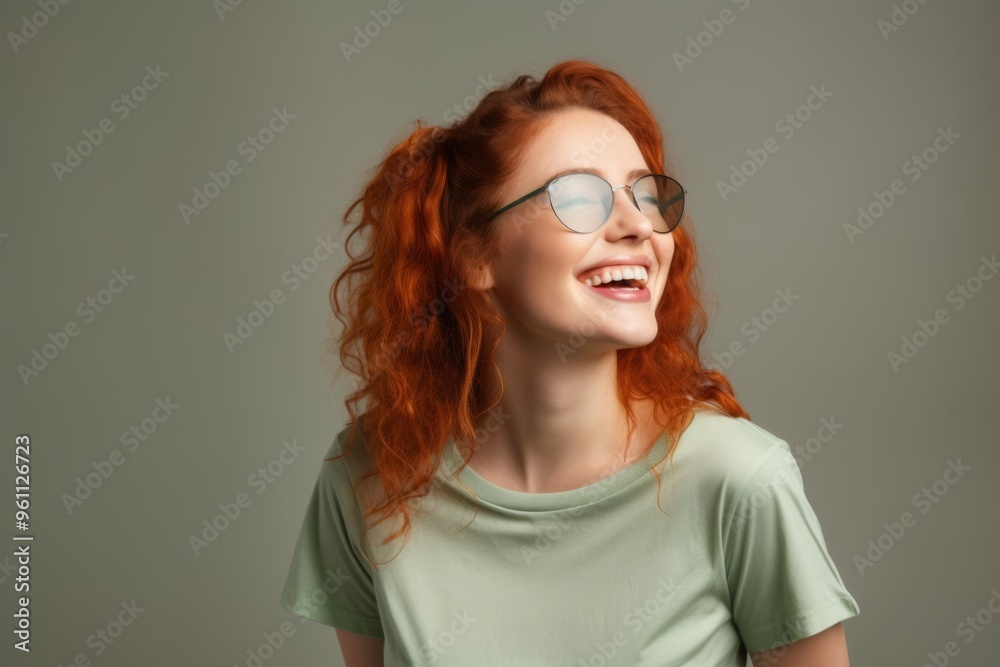Young woman with green eye patches on her face, isolated against a beige background. Close-up portrait of a ginger-haired girl wearing a T-shirt and looking away while smiling, applying a cosmetic gel
