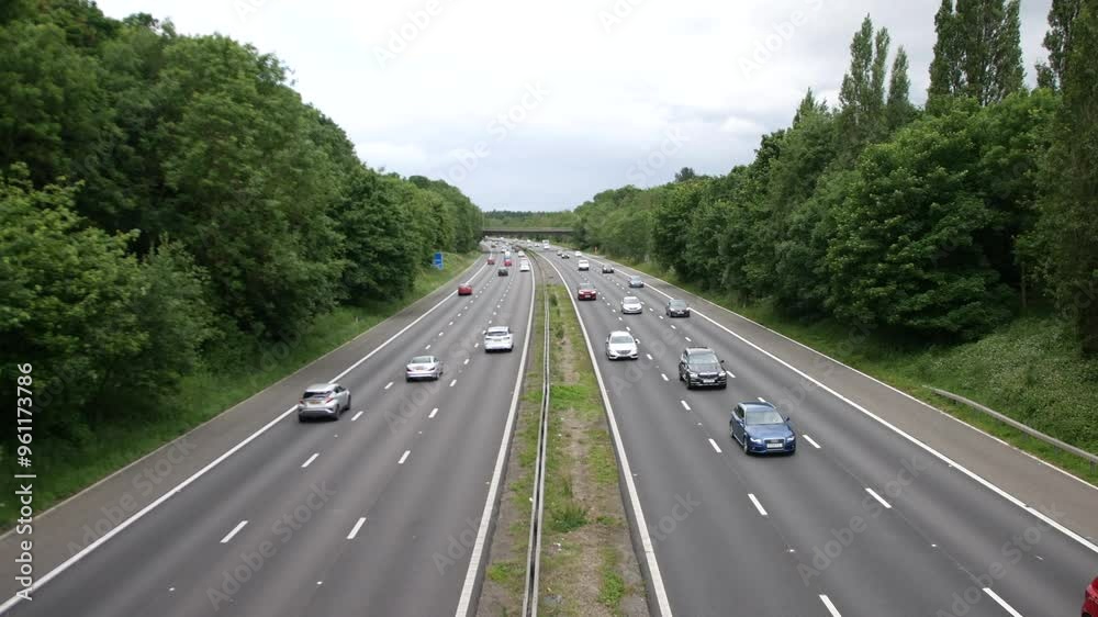 Cars travelling along the M1 motorway in England, surrounded by dense green trees on either side. The multi-lane highway shows light traffic under an overcast sky. Cars travelling along the M1 motorway in England, surrounded by dense green trees on either side. The multi-lane highway shows light traffic under an overcast sky.