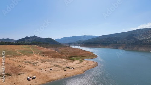 Aerial flies above Valdearenas beach and the dazzling Iznajar lake towards Puente Agroman bridge, on a spectacular hot Autumn day under azure blue skies. Cordoba province Spain.