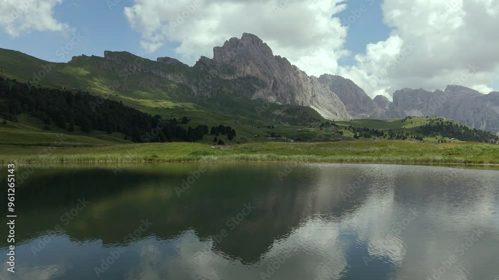 A tranquil alpine lake reflects the majestic Dolomites while a cyclist rides through a lush green meadow, under a sky dotted with clouds. A tranquil alpine lake reflects the majestic Dolomites while a cyclist rides through a lush green meadow, under a sky dotted with clouds.