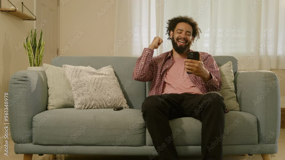 A joyful young African American man smiles while browsing the internet on his cellphone, seated on a soft sofa, reflecting a harmonious blend of relaxation and technology in his life A joyful young African American man smiles while browsing the internet on his cellphone, seated on a soft sofa, reflecting a harmonious blend of relaxation and technology in his life