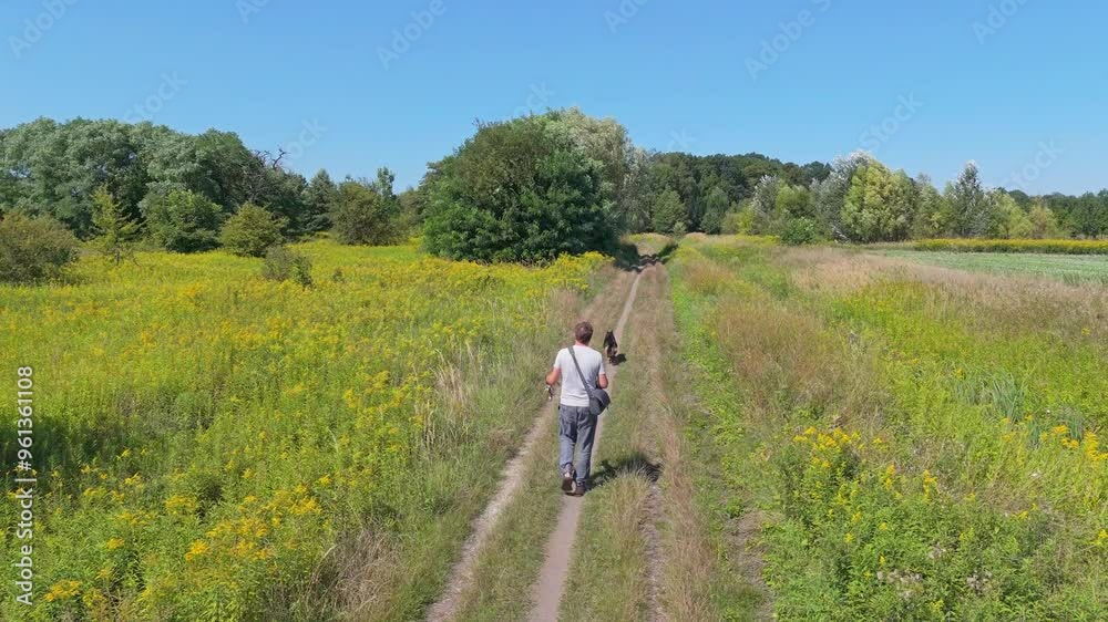 A man in linen pants and a light shirt strolls with his dog through fields and trees. Carrying a bag, he relaxes on a sunny day under a clear sky. A man in linen pants and a light shirt strolls with his dog through fields and trees. Carrying a bag, he relaxes on a sunny day under a clear sky.