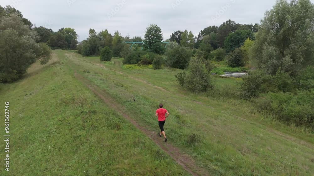 Running in the rain, a man in a red shirt and black track pants follows a narrow path along flood barriers outside the city. Slow-motion footage captures his outdoor fitness routine. Running in the rain, a man in a red shirt and black track pants follows a narrow path along flood barriers outside the city. Slow-motion footage captures his outdoor fitness routine.