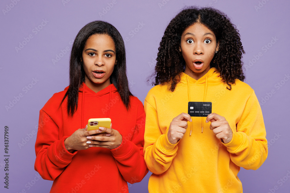 Two young amazed little kid teen girls women they wear red yellow hoodie casual clothes using mobile cell phone credit bank card shopping online isolated on plain purple background. Lifestyle concept. Two young amazed little kid teen girls women they wear red yellow hoodie casual clothes using mobile cell phone credit bank card shopping online isolated on plain purple background. Lifestyle concept.