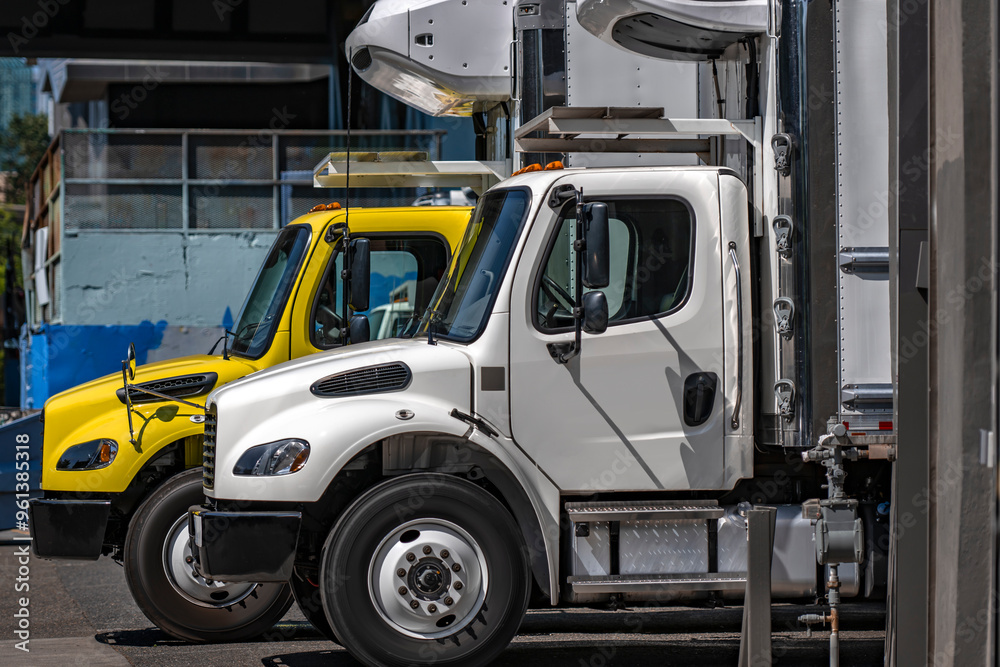 Two day cab middle duty rigs semi trucks with refrigerated box trailers standing side by side on the warehouse industrial parking lot Two day cab middle duty rigs semi trucks with refrigerated box trailers standing side by side on the warehouse industrial parking lot