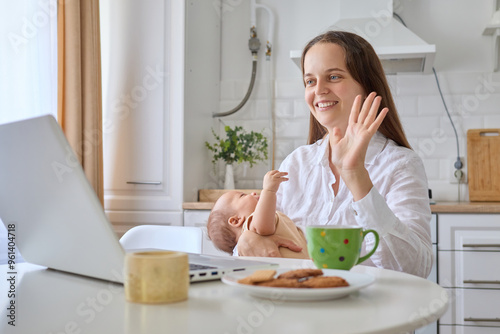 Cheerful smiling mother and baby in kitchen using technology for online communication mommy with newborn baby being happy and smiling woman waving hi gesture