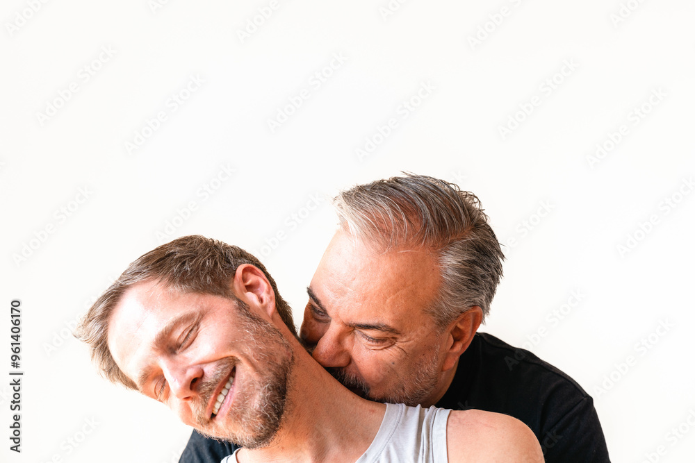 Fototapeta premium Älterer Mann mit grau schwarz meliert Bar, Haaren in schwarzem T-Shirt küsst einen jüngeren Mann, der vor ihm sitzt im grauen Tanktop mit Straßenköter blonden Haaren, Bart blaue Augen auf den Hals