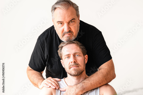 Älterer Mann mit grau schwarz meliert Bar, Haaren in schwarzem T-Shirt umarmt einen jüngeren Mann, der vor ihm sitzt im grauen Tanktop mit Straßenköter blonden Haaren, Bart blaue Augen photo