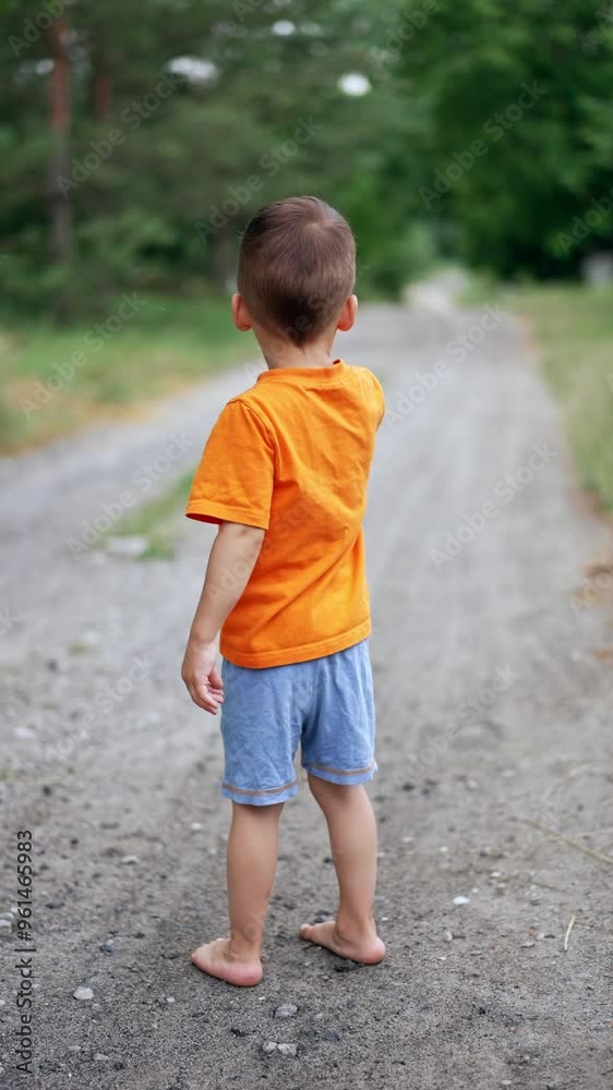 Caucasian toddler in orange t-shirt and blue shorts stands barefoot on the ground. A walk in the nature in summer. Vertical video. Caucasian toddler in orange t-shirt and blue shorts stands barefoot on the ground. A walk in the nature in summer. Vertical video.